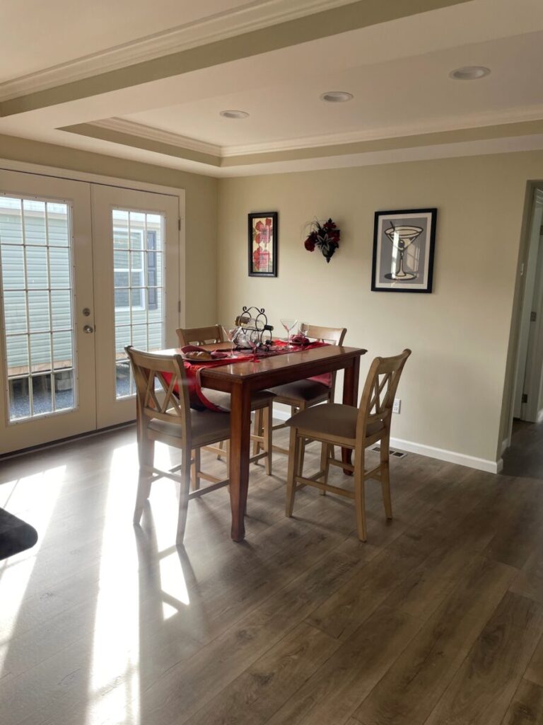Bright dining area with a wooden table set for four, red accents, and decorative pieces. Sunlight streams through French doors, adding warmth.
