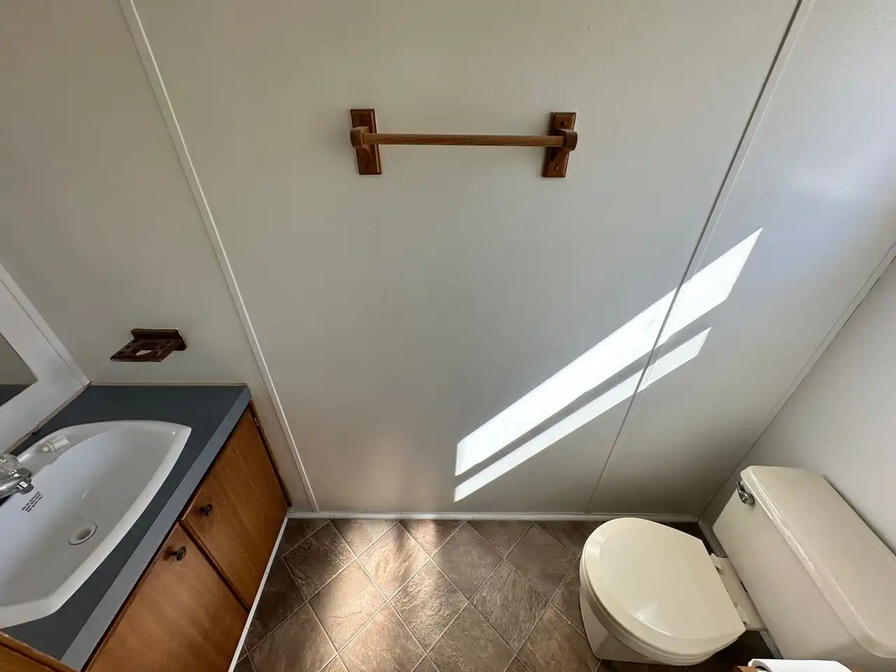 Small bathroom with a white toilet and sink on a dark countertop. Wooden towel rack on a white wall, diagonal sunlight creates a calm atmosphere.