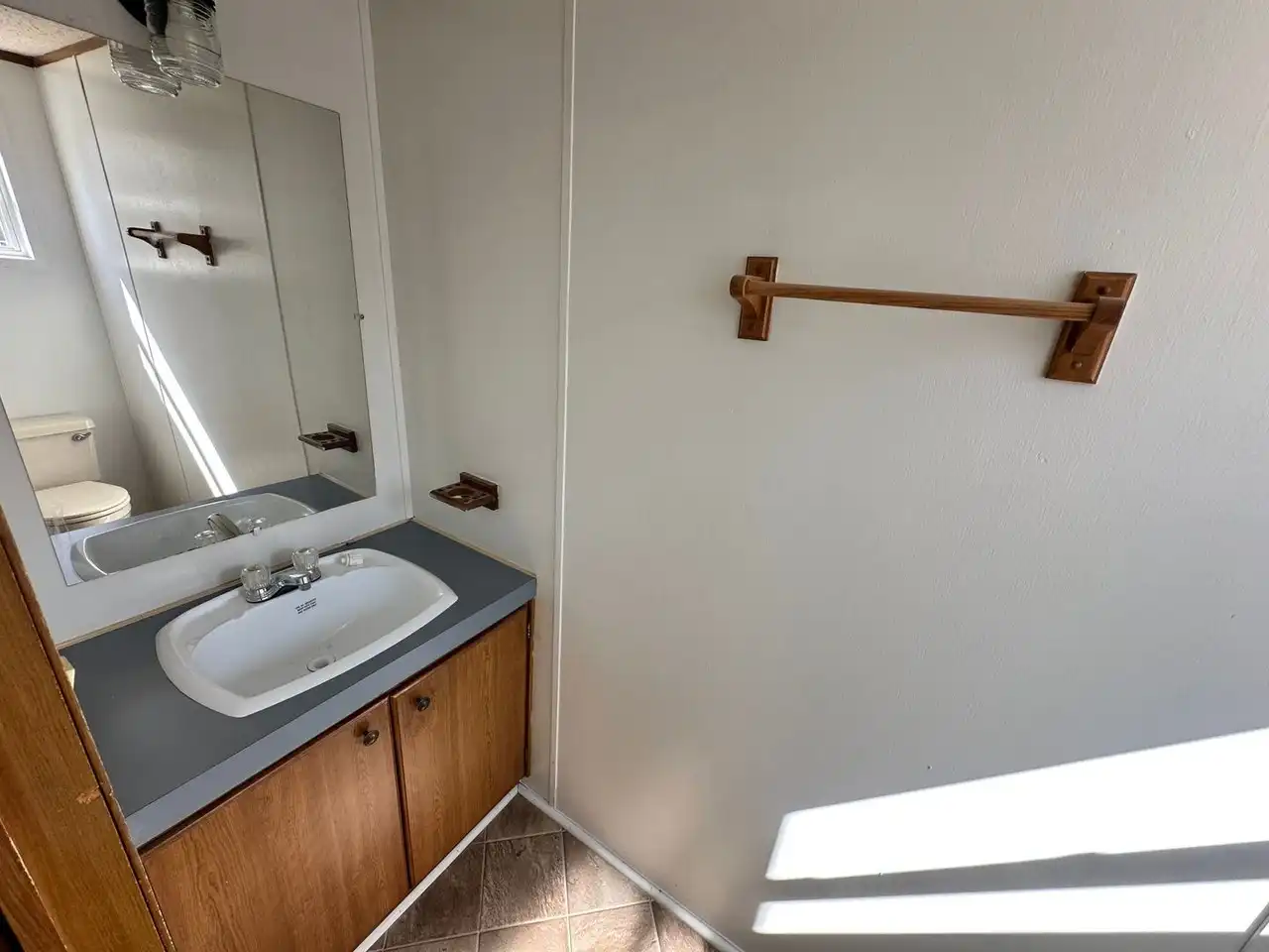 Small bathroom with a wooden vanity and white sink, reflecting in a mirror. A wooden towel bar is mounted on a plain wall, creating a minimalist look.
