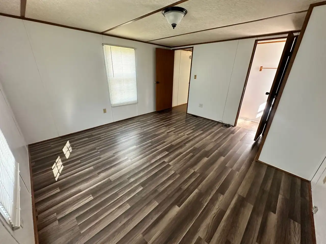 Empty room with white walls, dark wood flooring, and a ceiling light. A window with blinds lets in soft light. An open door leads to a small room.