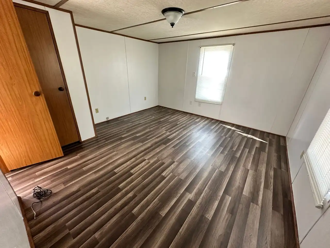 Empty room with white walls, dark wood laminate flooring, and a small window letting in light. A closed wooden door is in the corner, beside a ceiling light fixture, creating a calm, neutral tone.