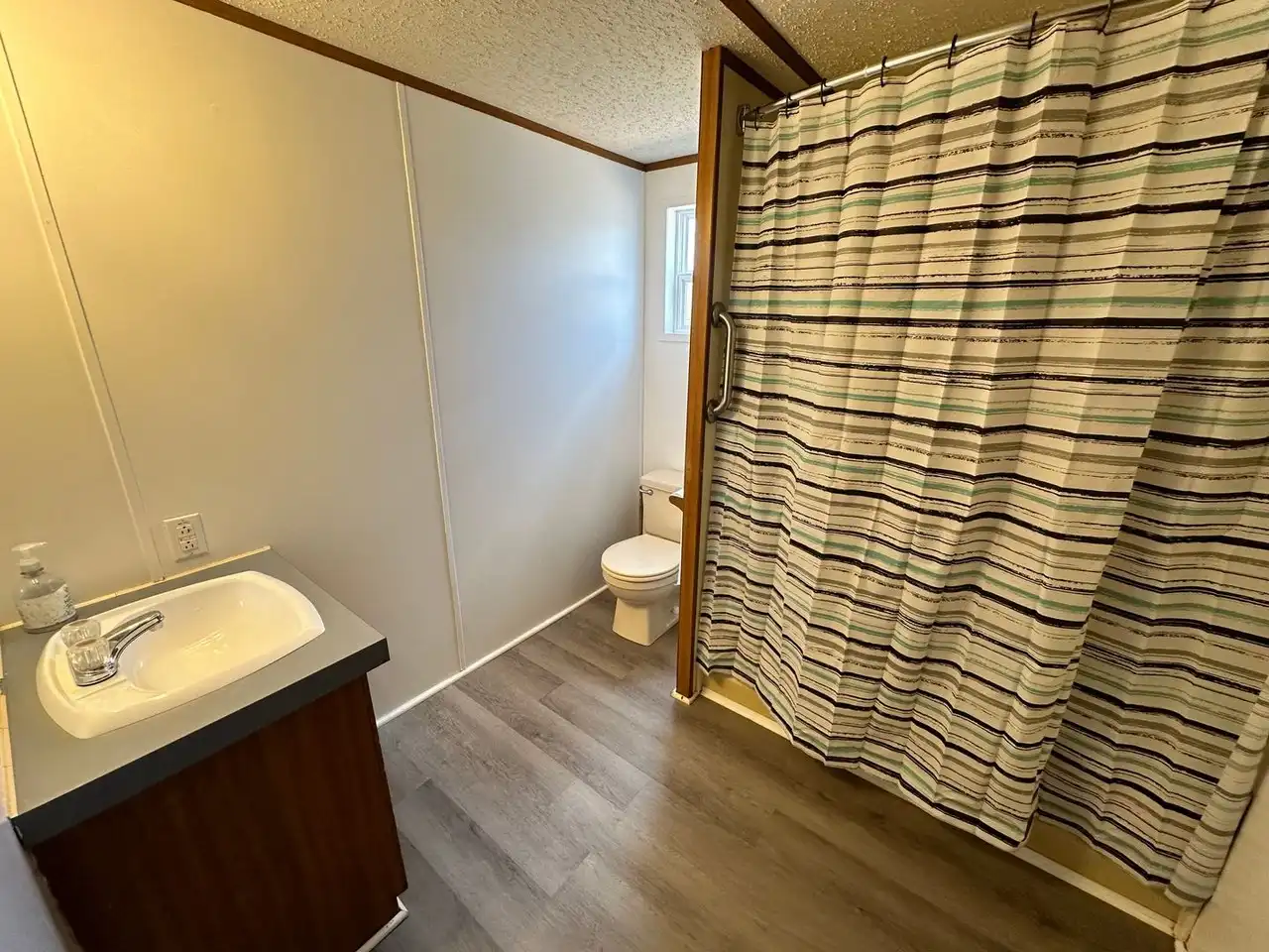 A compact bathroom with a striped shower curtain, a white toilet by a window, a modern sink with soap dispenser, and wood-effect flooring.
