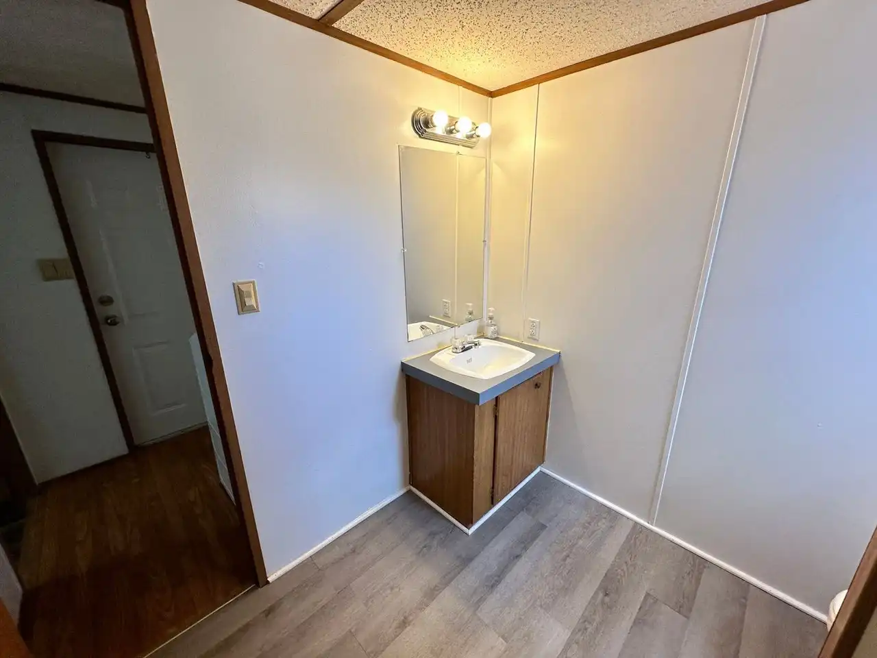 Simple bathroom with a small wooden vanity, a white sink, and a rectangular mirror. Bright overhead lighting highlights light-colored walls and vinyl flooring.
