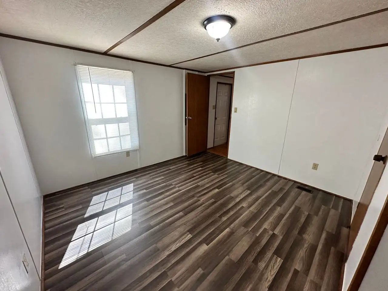 Empty room with white walls, dark wood laminate flooring, and a ceiling light. A window with blinds lets sunlight in, creating a calm atmosphere.