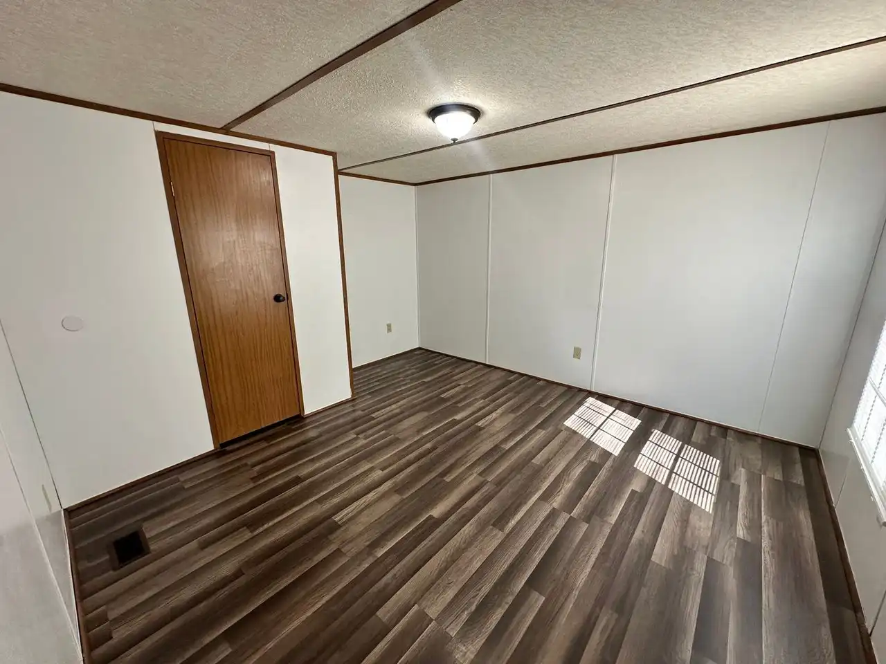 An empty room with white walls, brown wood laminate flooring, and a wooden door. Natural light streams through a window, creating a calm atmosphere.