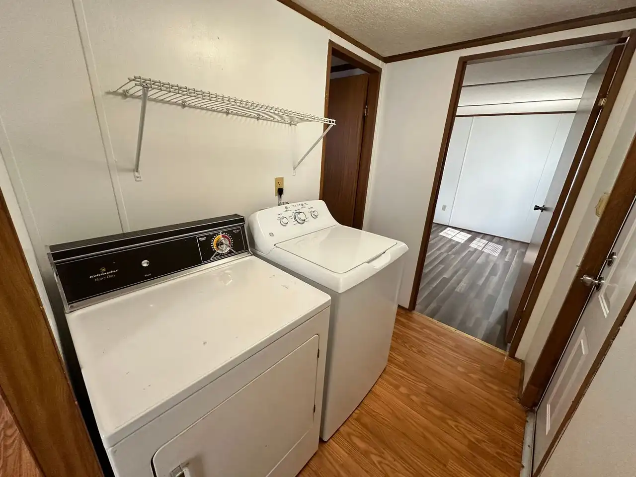 Compact laundry area with a classic washer and dryer on a wooden floor, a wire shelf above. Leads to a bright, empty room with laminate flooring. Cozy atmosphere.