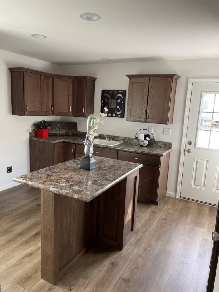A modern kitchen with dark wood cabinets, marble countertops, a central island with a vase and flowers, wooden flooring, and a white door.