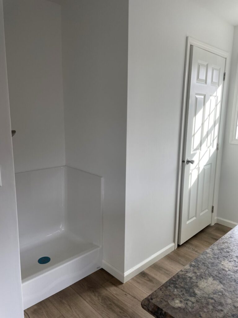 A bright room featuring a corner shower with a white base, a closed white door, and wooden flooring. Sunlight casts shadows on the door, creating a serene ambiance.