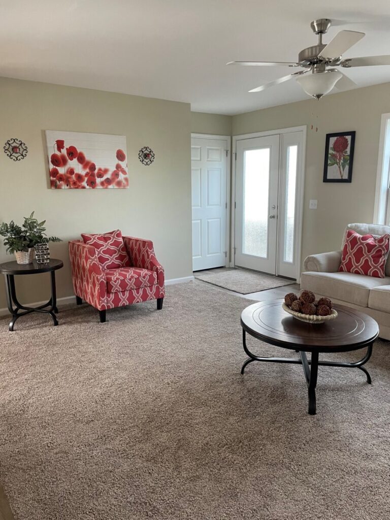 Cozy living room with red-patterned armchair and pillows, beige carpet, round wooden coffee table, floral wall art, and large windows creating a bright, welcoming atmosphere.
