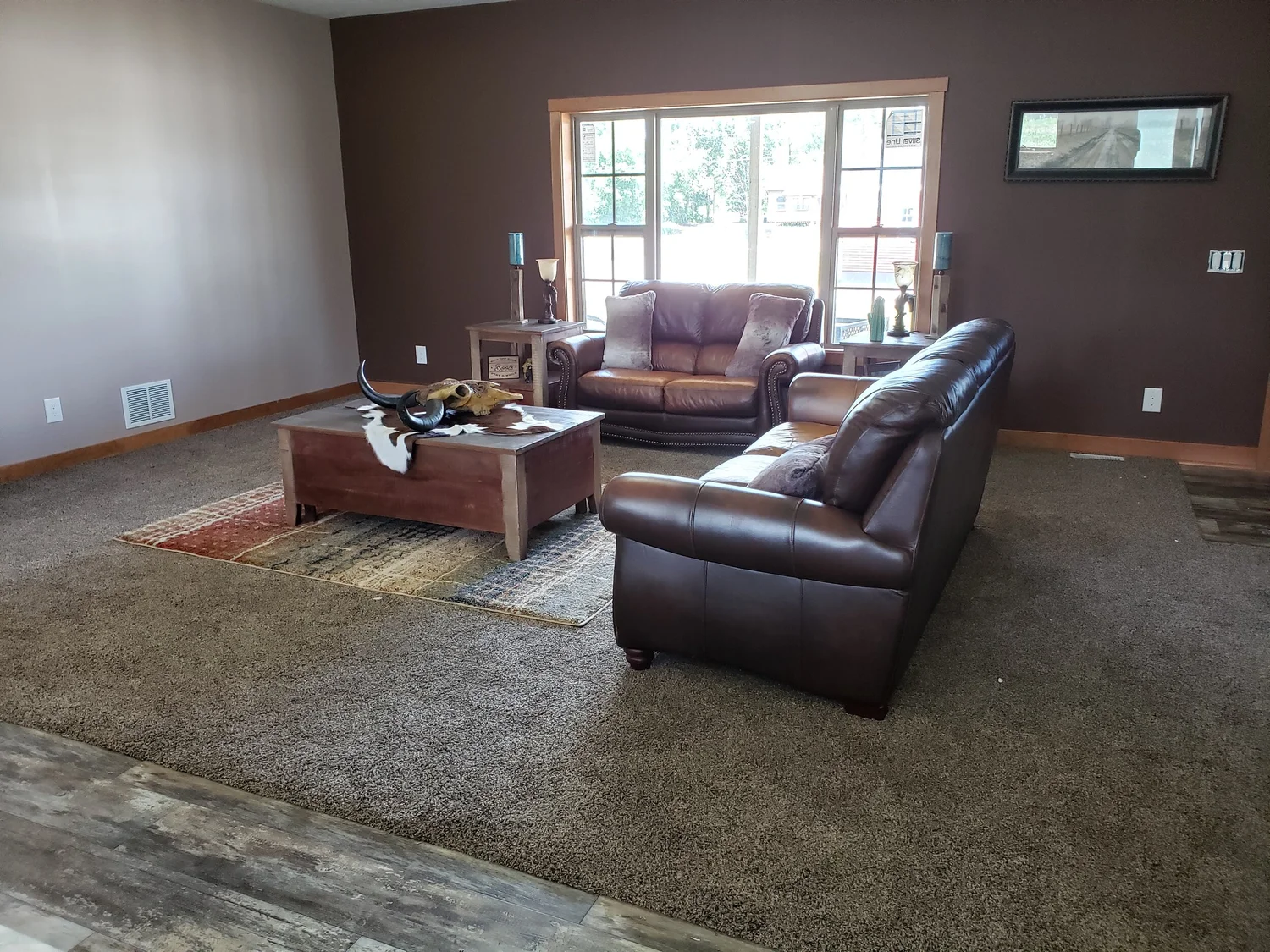 A cozy living room with brown leather sofas and a wooden coffee table on a patterned rug. Sunlight streams through a large window, creating a warm ambiance.
