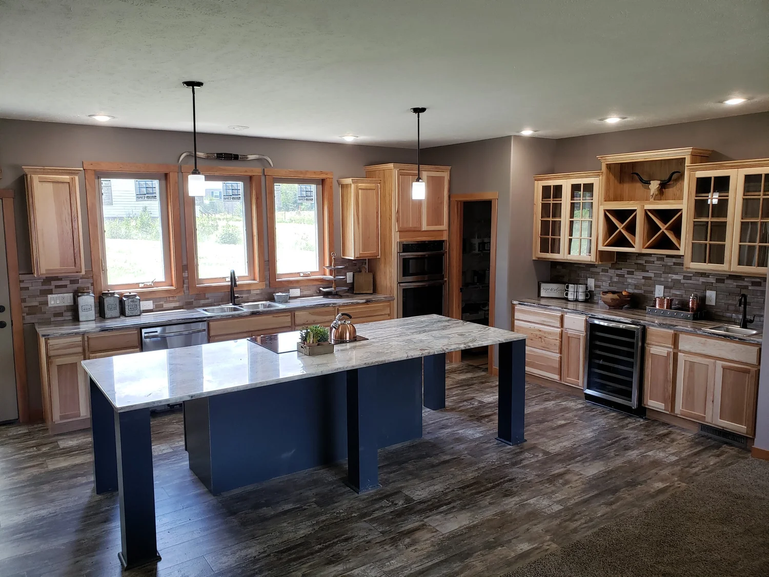 Spacious kitchen with wood cabinets, marble countertops, and a central island. Pendant lights hang above, accented by natural light from large windows.