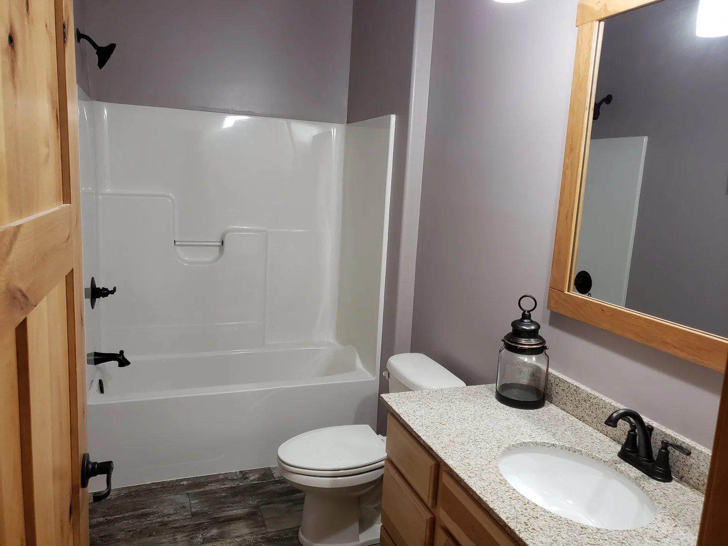 Compact bathroom with a white bathtub, shower, and toilet. Gray walls, wood-framed mirror, granite countertop, and black fixtures. Clean and modern.
