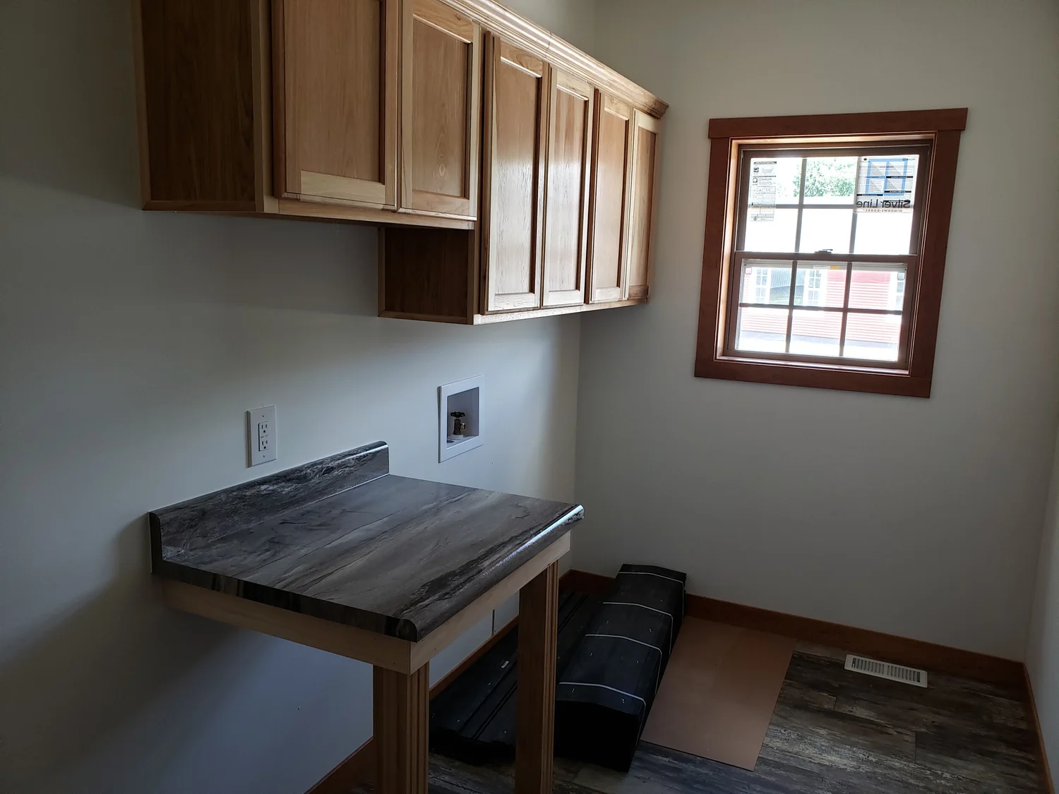 A small laundry room with light wooden cabinets, a marble countertop, and a window with wooden trim. The room is well-lit and organized.