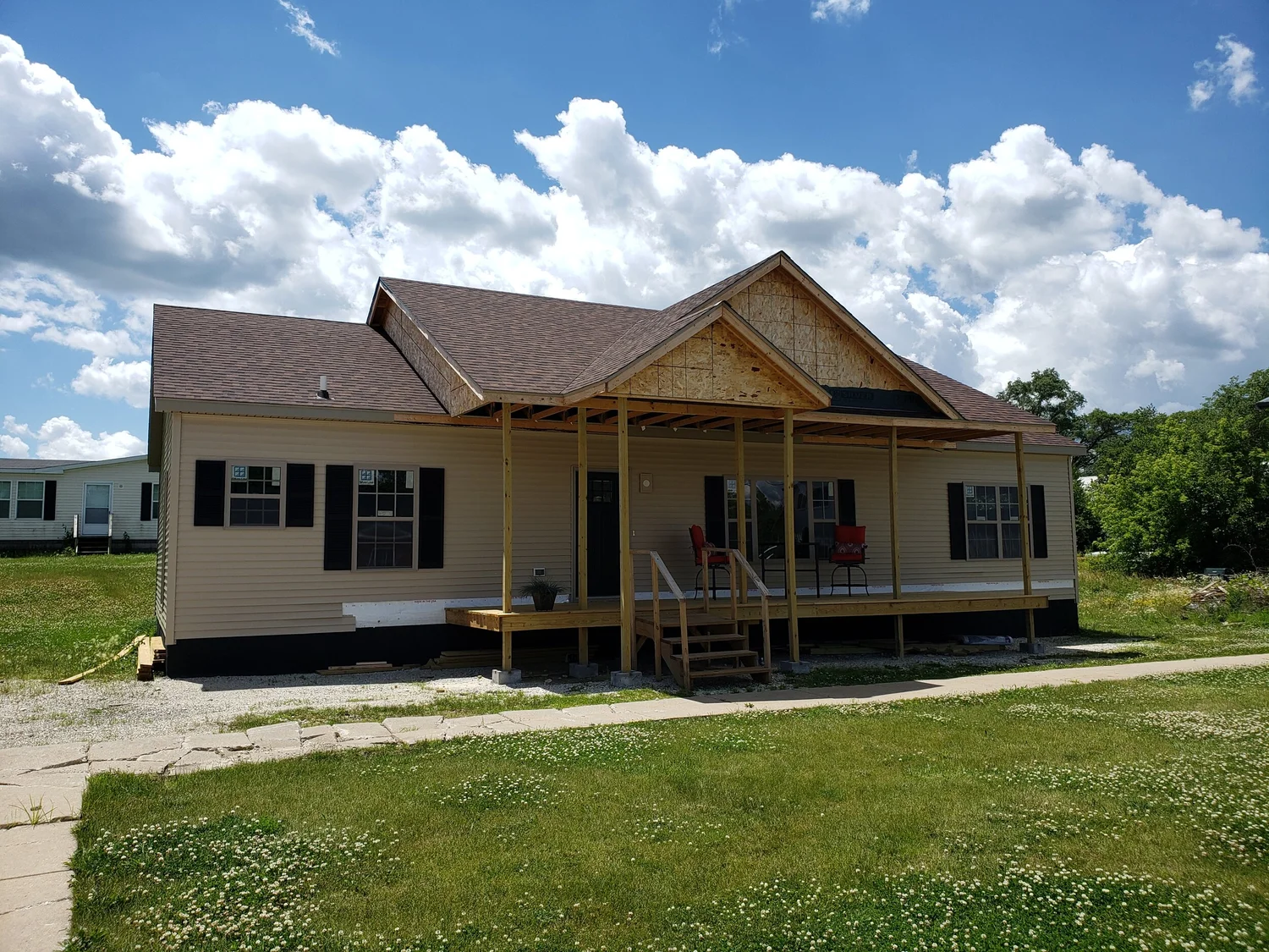 Single-story house with beige siding and a partially constructed wooden porch under a sunny, blue sky with scattered clouds. Lawn surrounds the home.