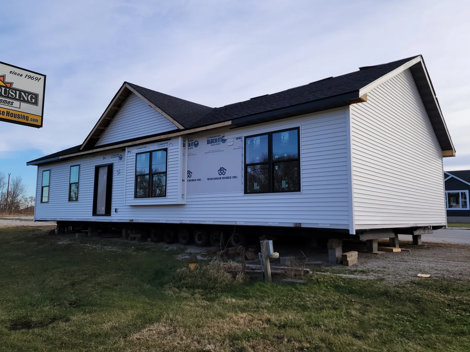 A white modular home partially constructed on a gravel lot, with exposed wheelbase beneath. A sign for a housing company is seen in the background.