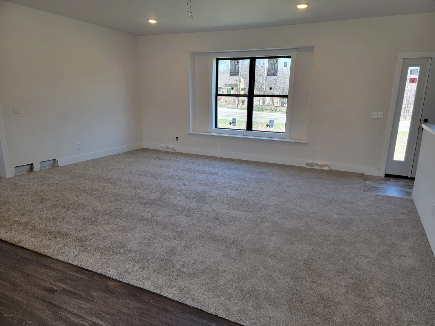 Spacious room with beige carpet and wooden floor, featuring a large window and glass door. Bright natural light creates a calm atmosphere.