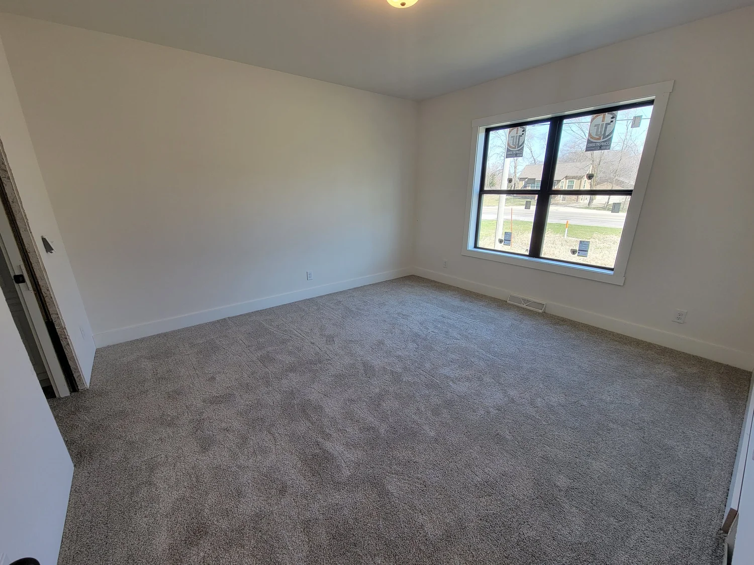 Empty room with gray carpet, white walls, and large window. Natural light streams in, creating a calm, spacious atmosphere. Open doorway on the left.