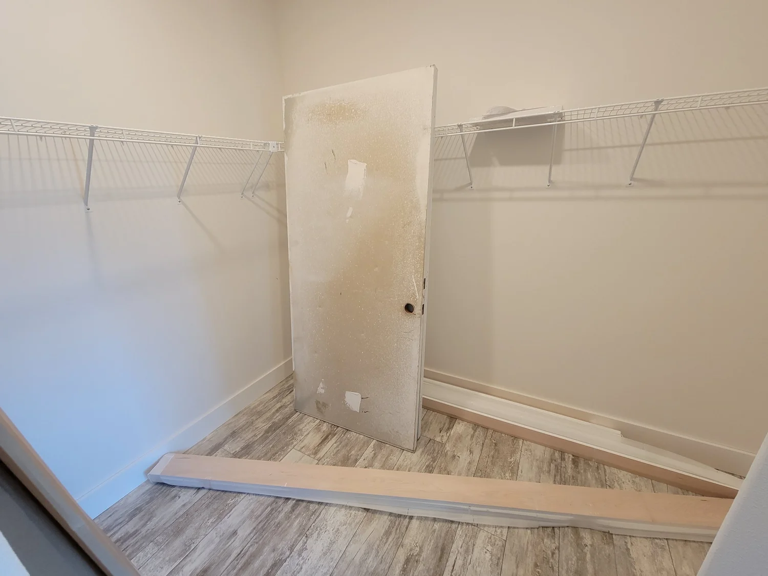 Empty closet with light wooden floor, beige walls, and three white wire shelves. A dirty, removed door and wooden boards are on the floor, suggesting renovation.