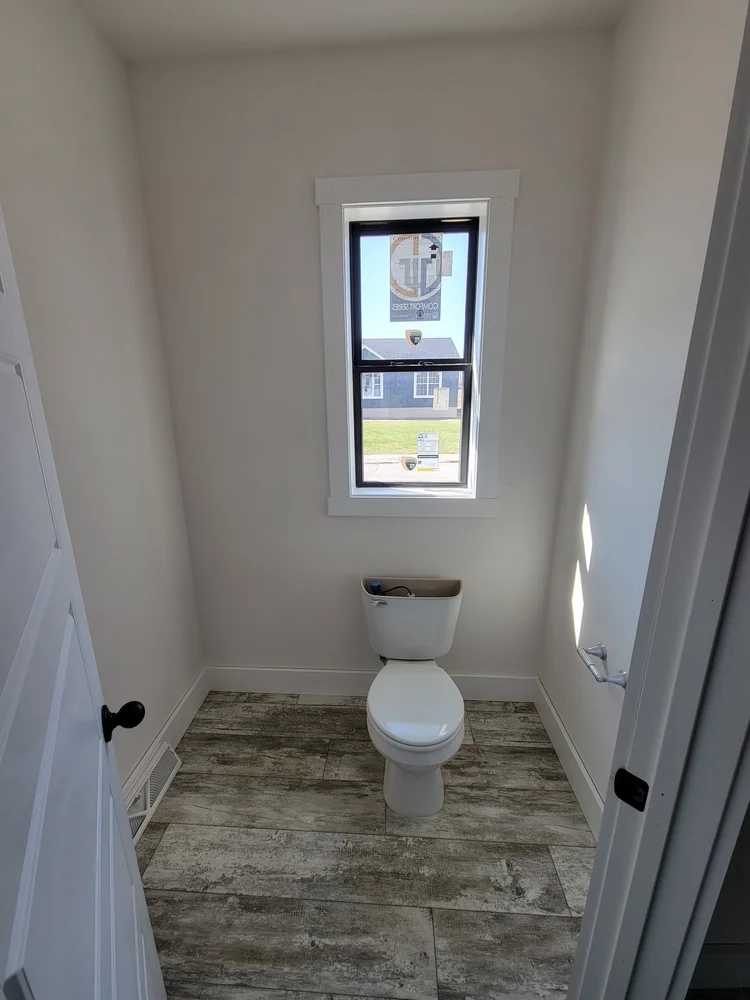Small, bright bathroom with a single toilet, wood-look tile floor, and a window offering natural light. The clean, minimalistic space feels calm.