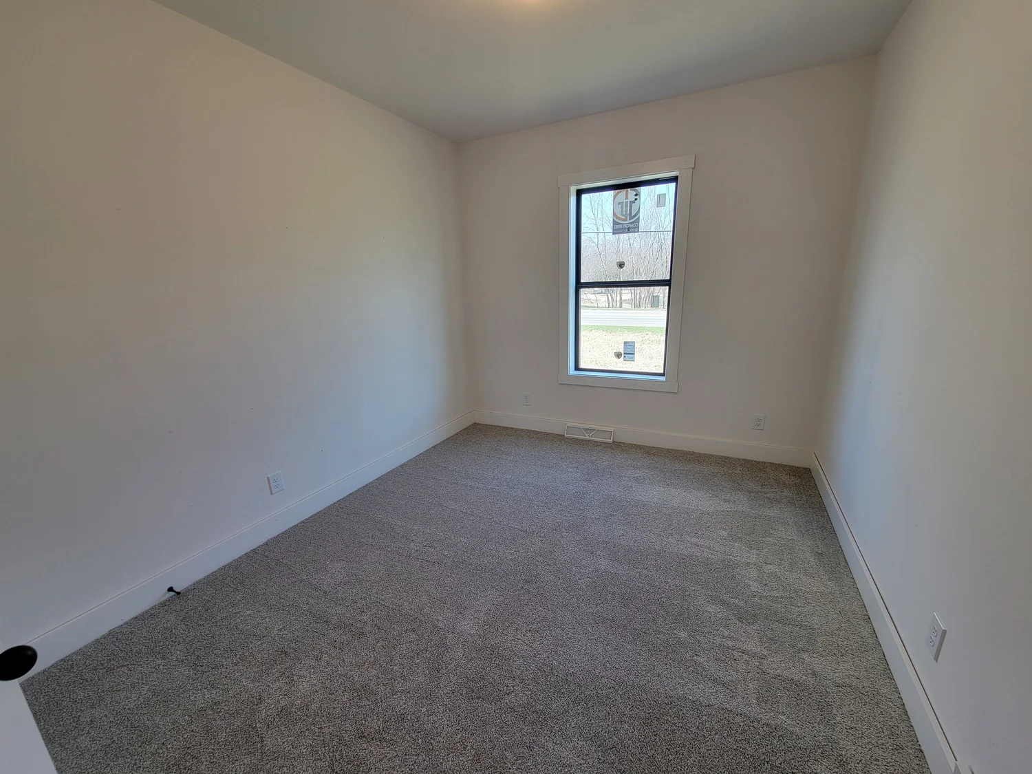 Empty room with white walls and gray carpet, featuring a single window revealing a view of trees and grass outside. The space feels bright and minimalistic.