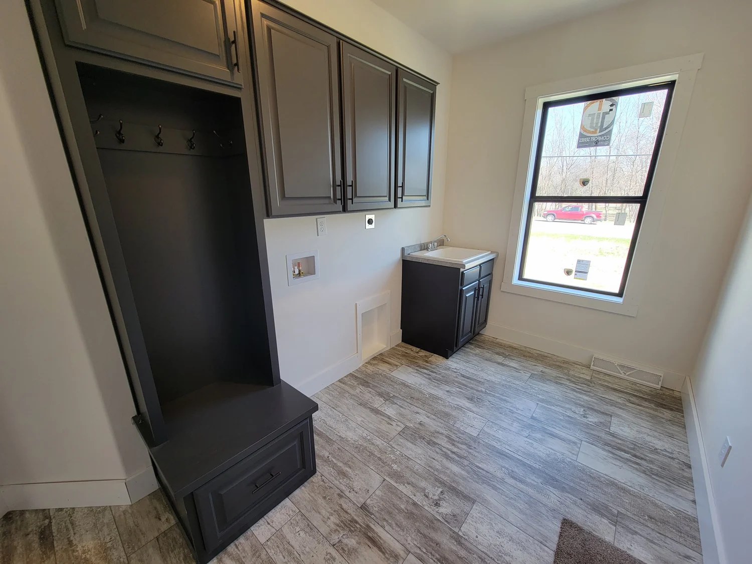 Modern laundry room with dark gray cabinets and hooks on the left, a small sink on the right, and a large window flooding the room with natural light.