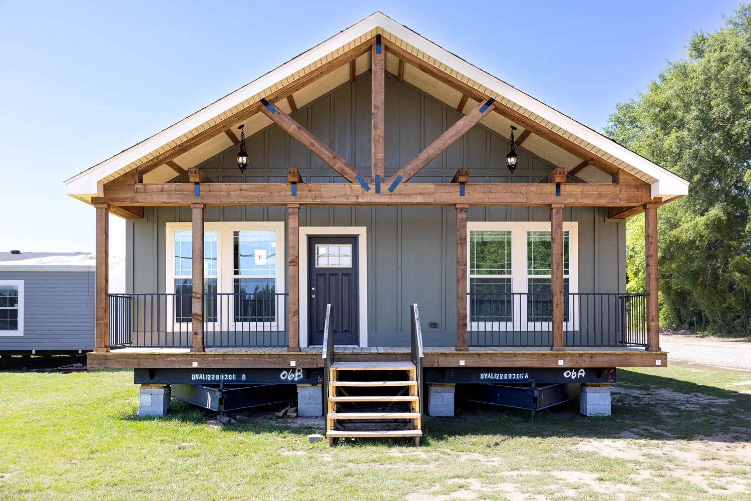 A small prefabricated house with a gabled roof and wooden beams, featuring a front porch with metal railings and stairs, set on grass under a clear blue sky.