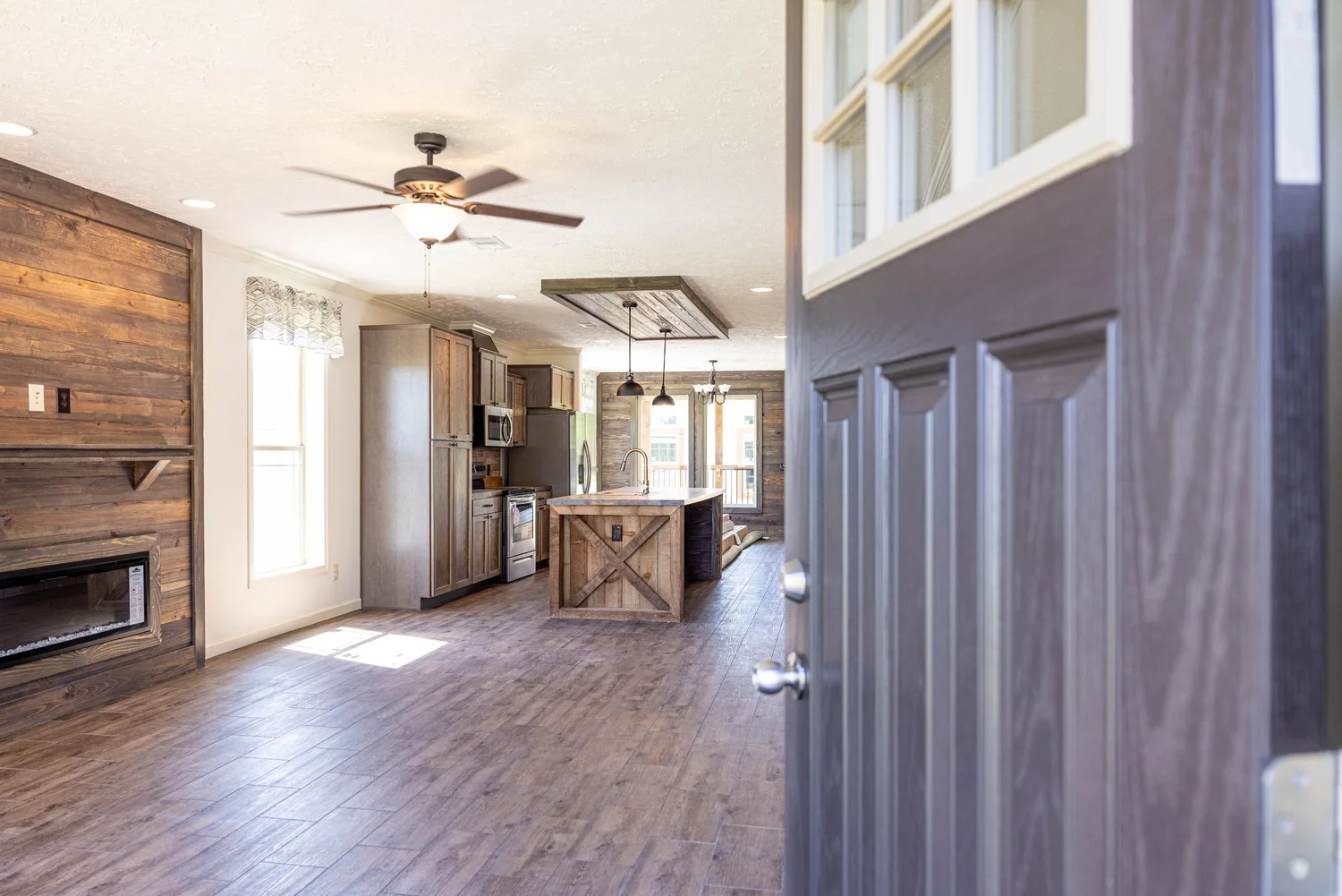 Open door to a rustic, modern kitchen with wood floors, exposed wood walls, an island, pendant lights, ceiling fan, and large windows, conveying warmth.
