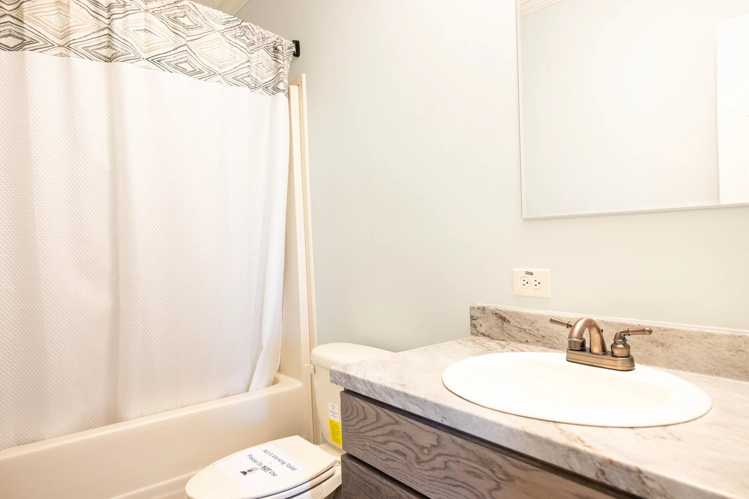 A clean bathroom with a white bathtub and patterned shower curtain. The sink, on a wooden vanity, has a marble countertop and bronze faucet, creating a fresh tone.