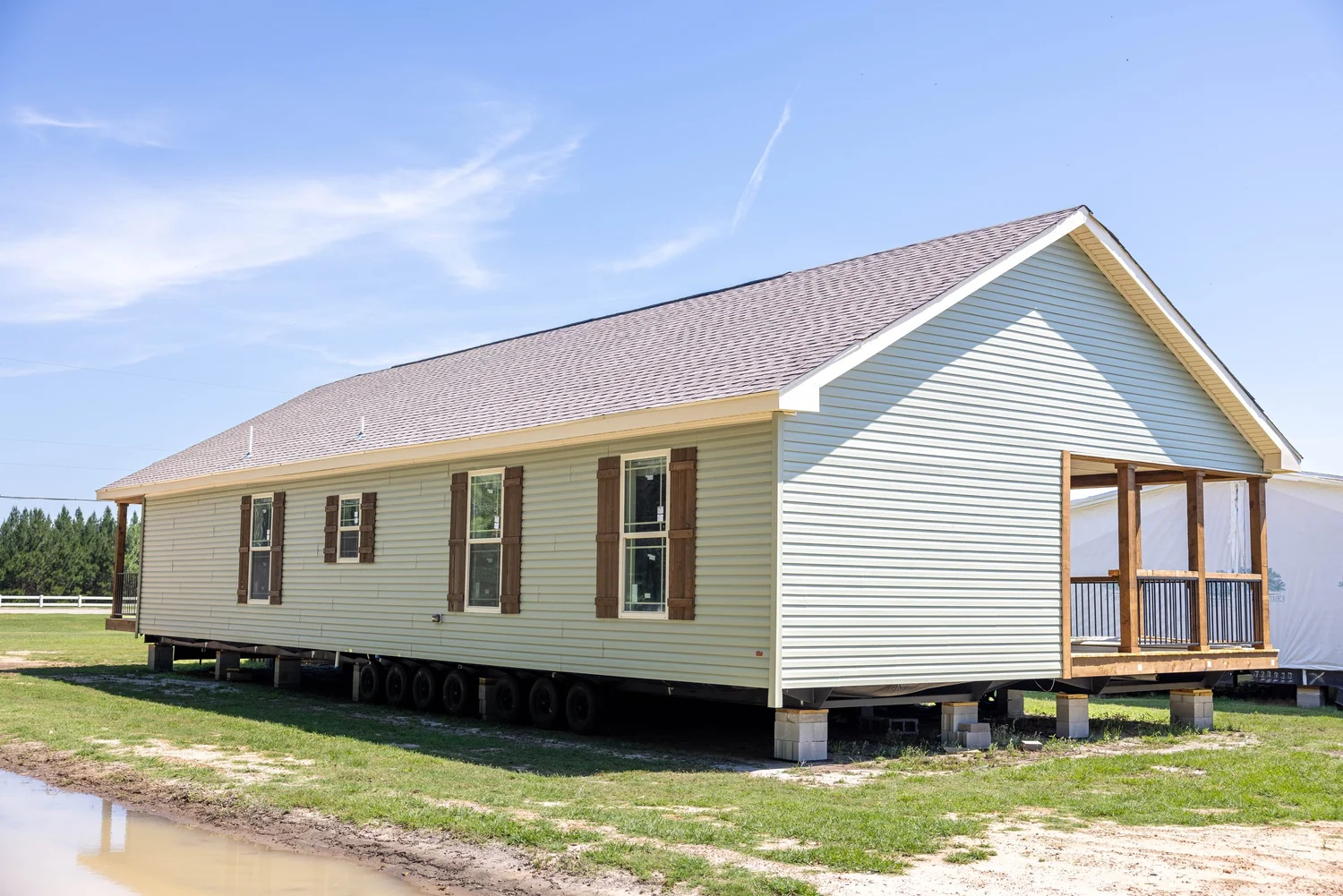A newly constructed modular home sits elevated on blocks in a grassy area. It features light siding, multiple windows, and a small porch. Sunny sky above.