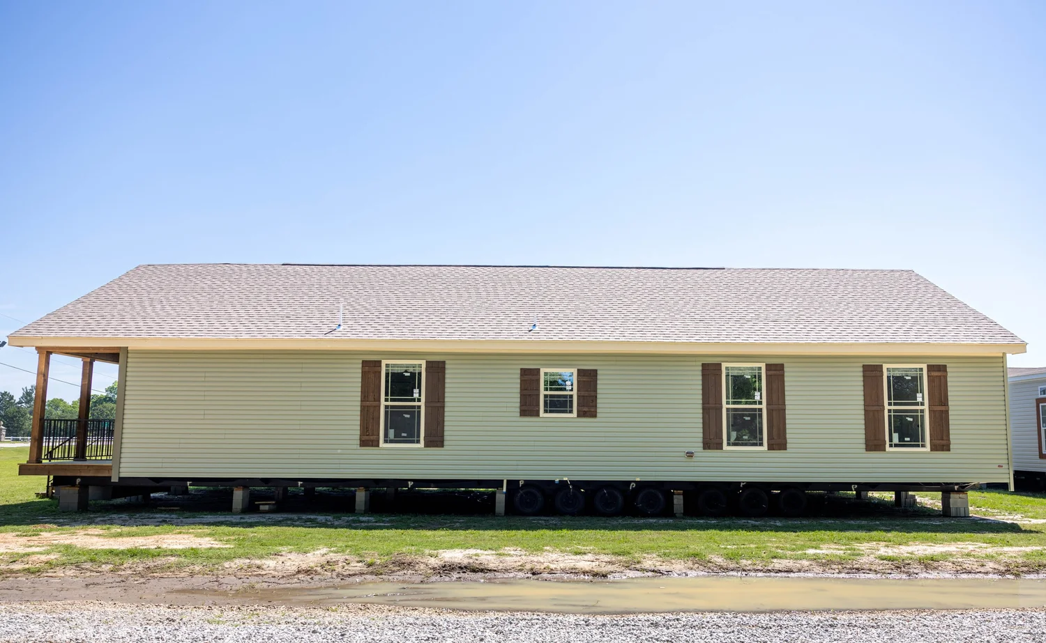 A beige manufactured home with brown shutters sits on raised supports in a grassy area under a clear blue sky, conveying a sense of simplicity and potential.