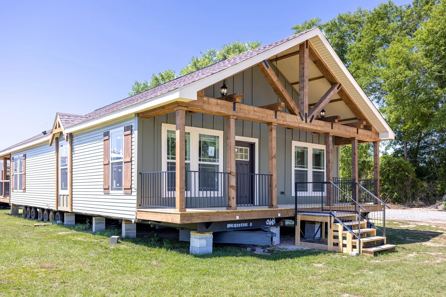 A modern prefabricated home with a wooden porch and gabled roof sits on a grassy lot. The exterior features gray siding and large windows.