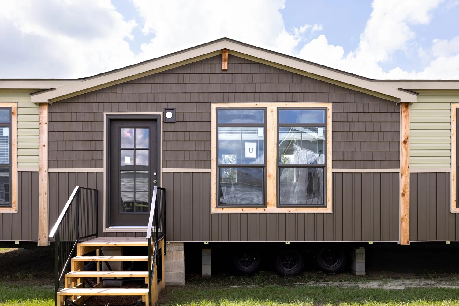 A modern prefab house with a dark brown exterior, two large windows, and front steps leading to a glass door. The sky is bright and partly cloudy.