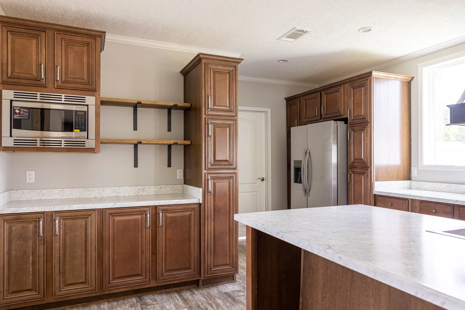 Spacious kitchen with brown wooden cabinets and marble countertops. Features a stainless steel fridge, built-in microwave, open shelves, and bright natural light.