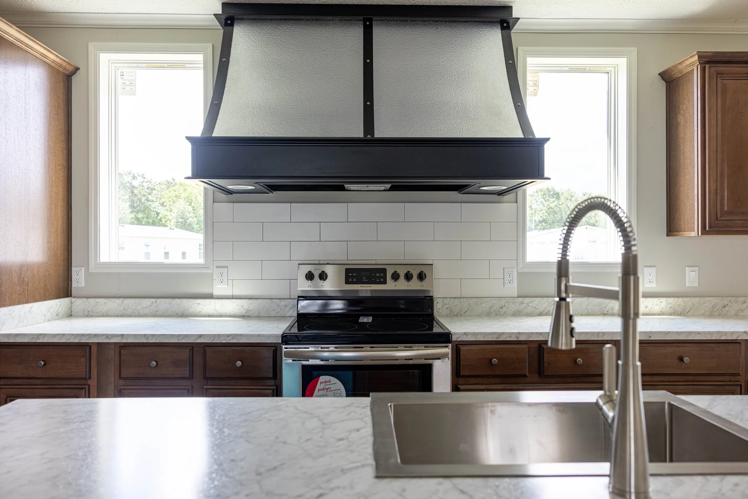 Modern kitchen with a large black range hood, stainless steel stove, white subway tiles, light marble countertops, and a silver faucet and sink.