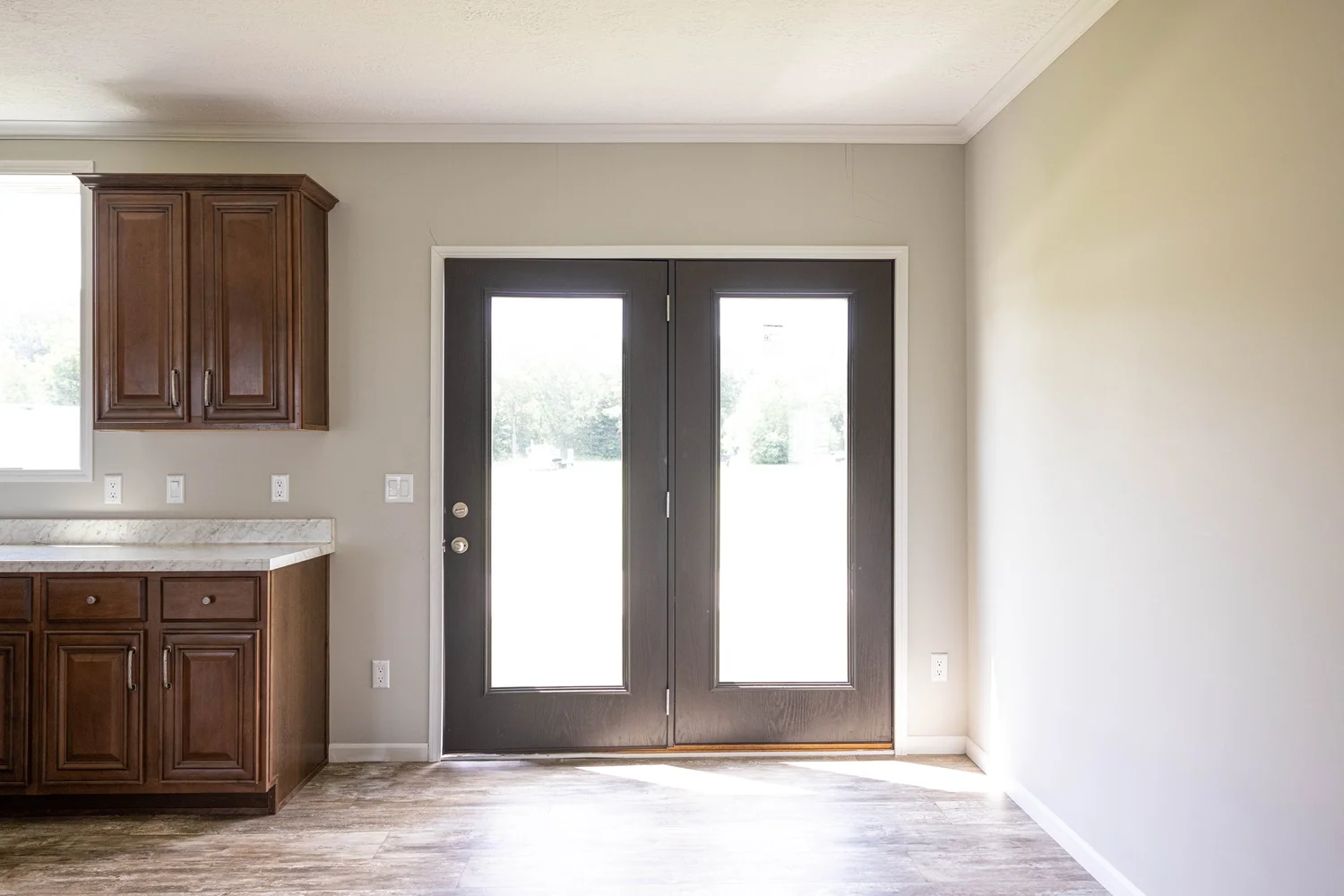 Bright room with dark double glass doors letting sunlight stream in. To the left, brown wooden cabinets and a countertop. Simple and inviting ambiance.