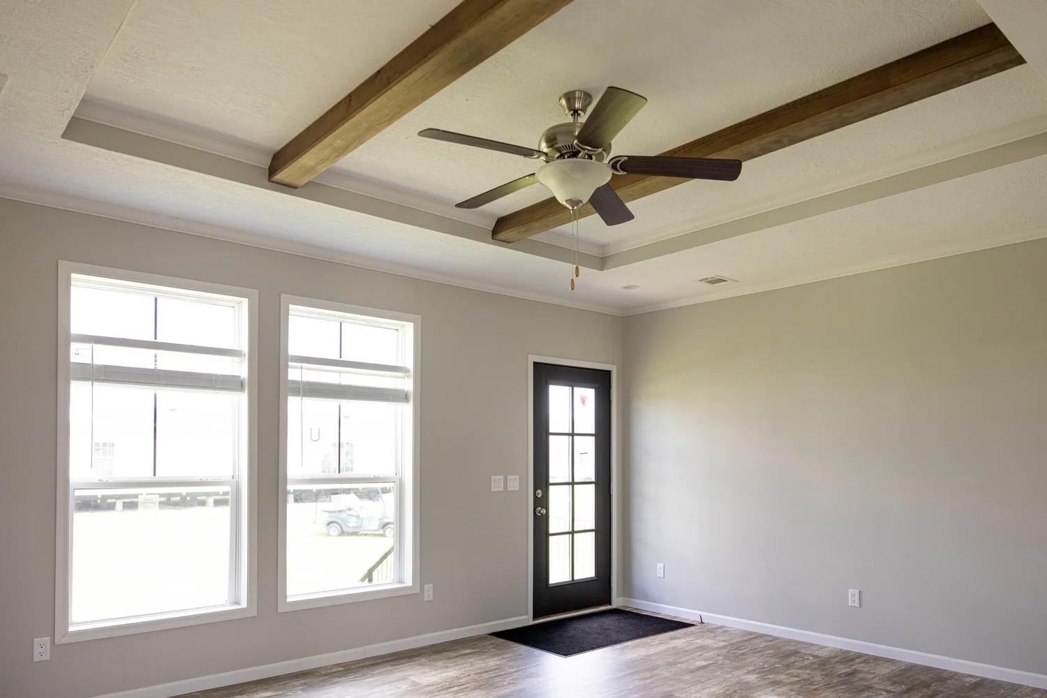 A minimalist room with light gray walls, wooden ceiling beams, a ceiling fan, two large windows, and a black-framed glass door; calm and airy ambiance.