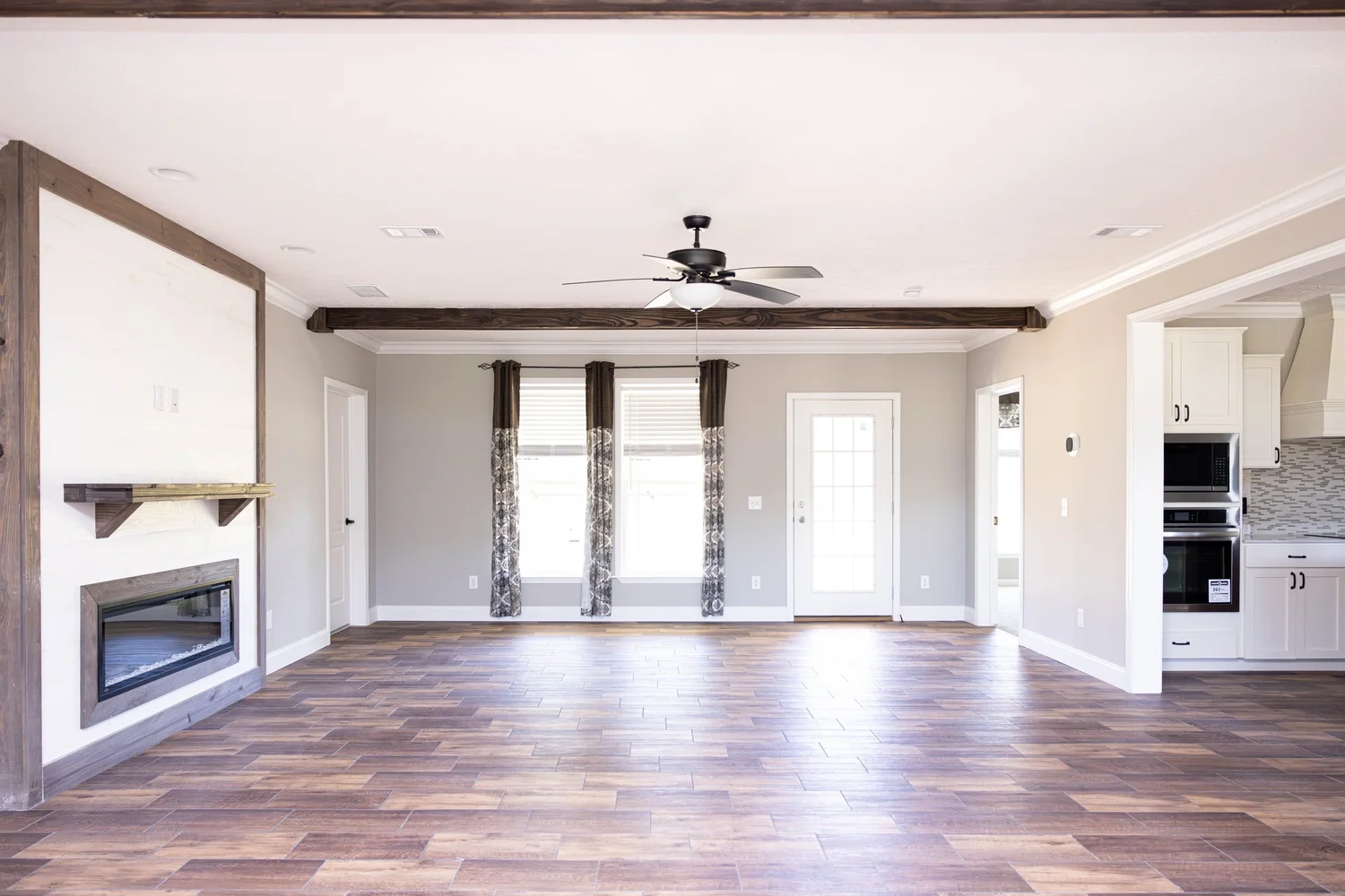 Spacious, modern living room with wooden floor, beige walls, and dark ceiling beams. Features a ceiling fan, fireplace, and adjacent kitchen. Bright and airy.