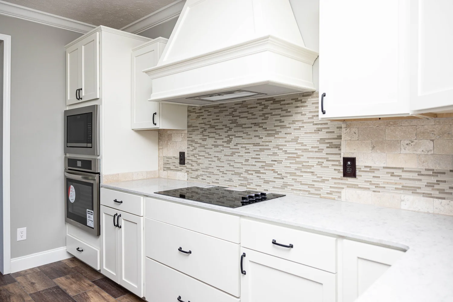 Modern kitchen with white cabinets, a sleek black cooktop, and stainless steel appliances. Stone-tile backsplash adds texture. Bright, clean space.