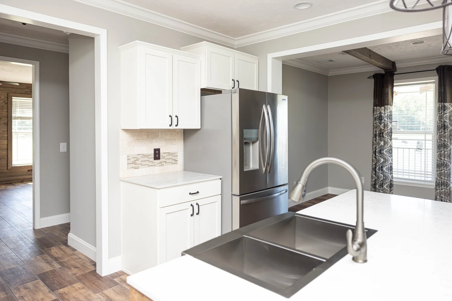 Modern kitchen with white cabinets, stainless steel fridge, and a sleek faucet. Wooden floor, gray walls, and large window create a bright, airy feel.