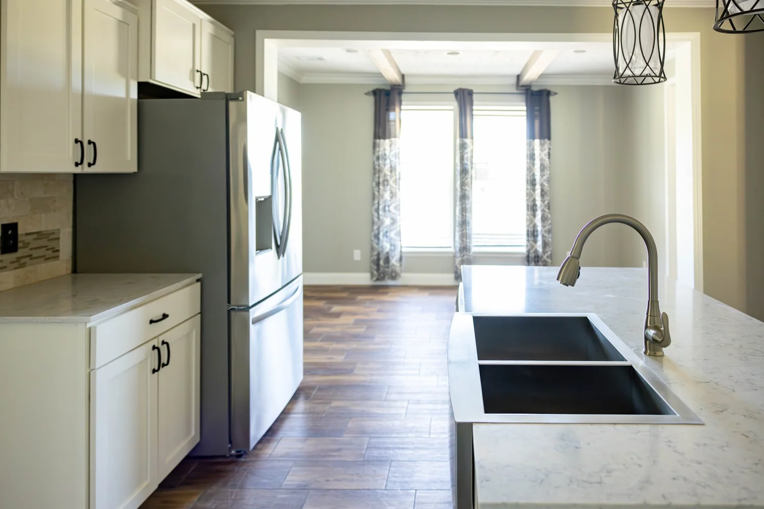 Modern kitchen with white cabinets, stainless steel fridge, dual sink on marble countertop, and wooden floor. Bright window and contemporary lighting.
