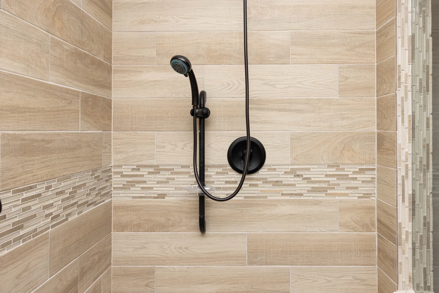 Modern shower with light wood-style tiles and a black handheld showerhead. Horizontal mosaic strip adds elegance. Clean, minimalist design.