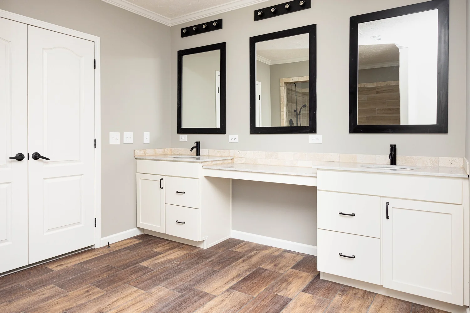 Spacious bathroom with dual white vanities, black-framed mirrors, and modern faucets. Wood-textured floor and soft lighting create a clean, elegant ambiance.