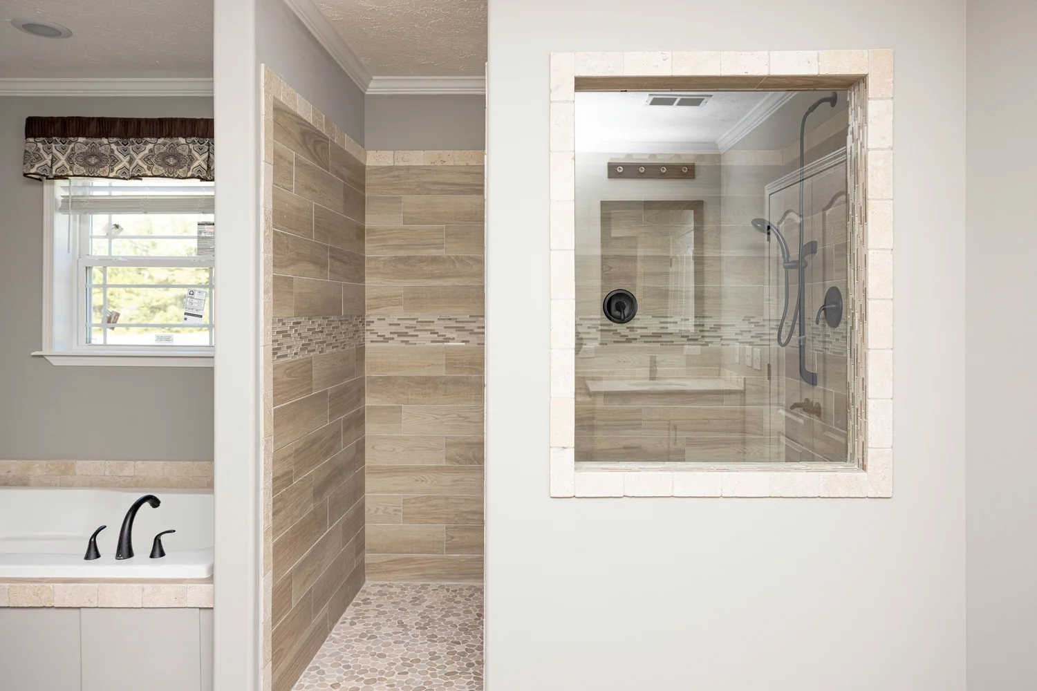 Modern bathroom with beige tile shower featuring a glass door, adjacent to a bathtub with black fixtures. Natural light from a small window.