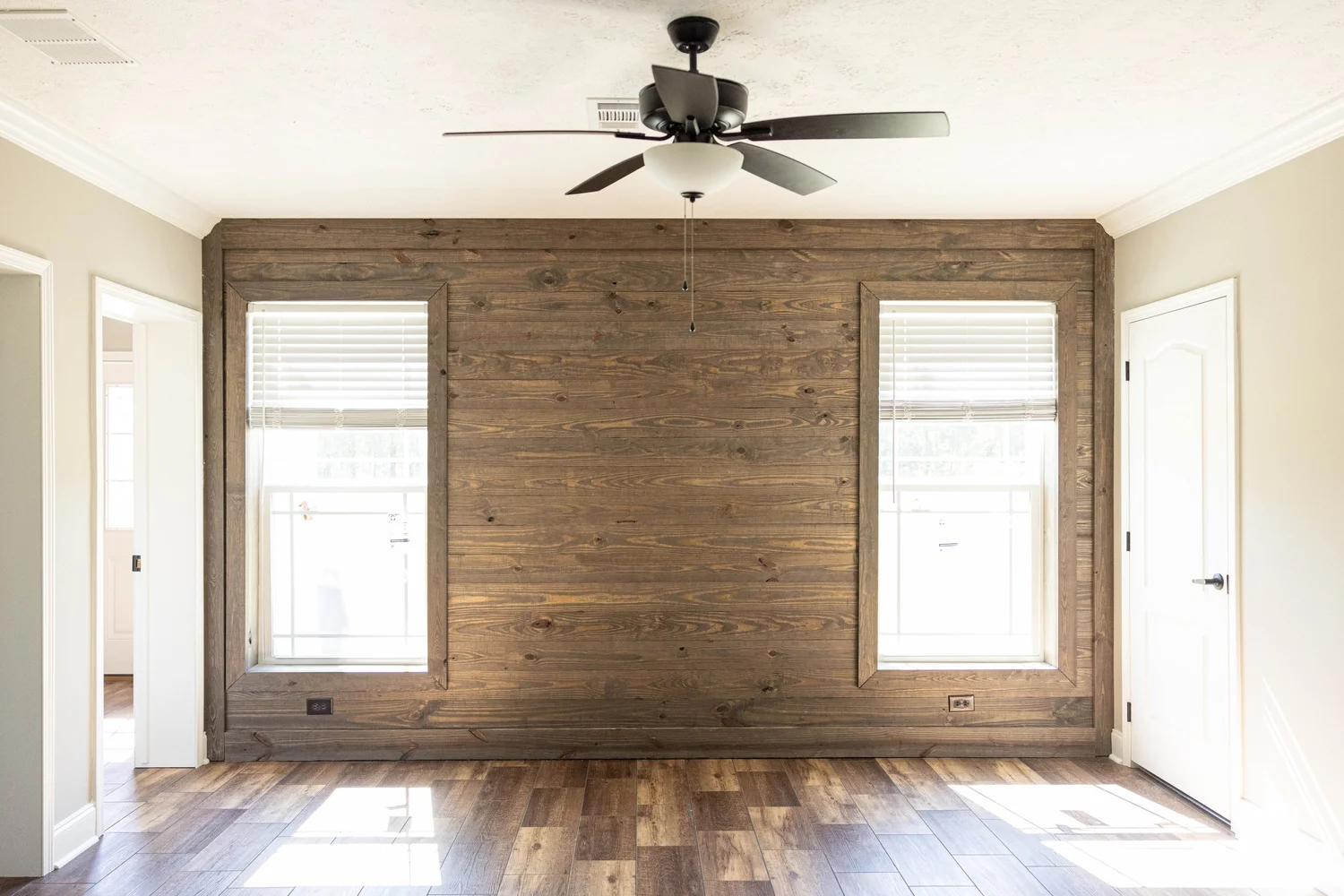 Room with a wood accent wall featuring two windows, white blinds, and a ceiling fan. The warm tones of the wood floor add a cozy feel.