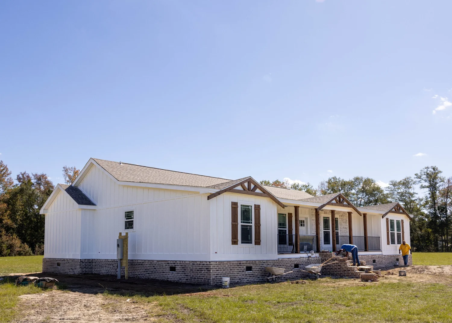 Large white house with a brick foundation on a grassy lawn, under a clear blue sky. Workers are building steps, creating a calm and constructive atmosphere.