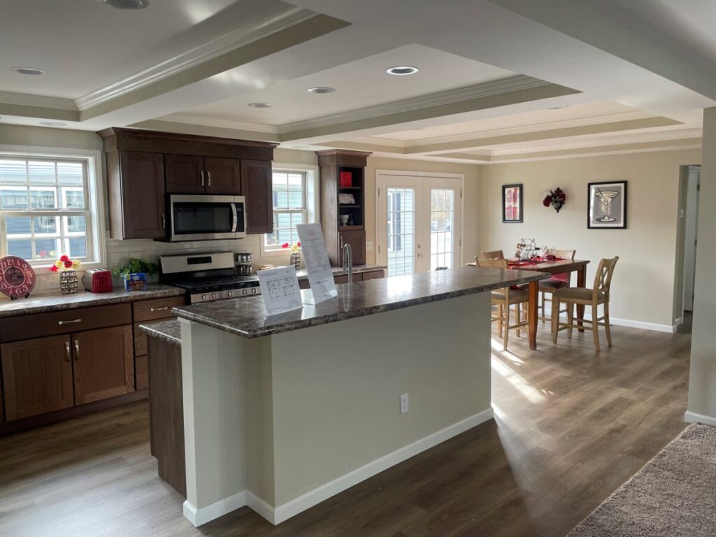 A modern kitchen with brown cabinets and stainless steel appliances opens to a dining area with a table set for four. Sunlight streams through glass doors.