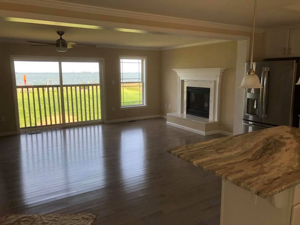 Spacious living room with large windows overlooking a serene lake. The room features a ceiling fan, hardwood floors, a marble countertop, and a white fireplace.
