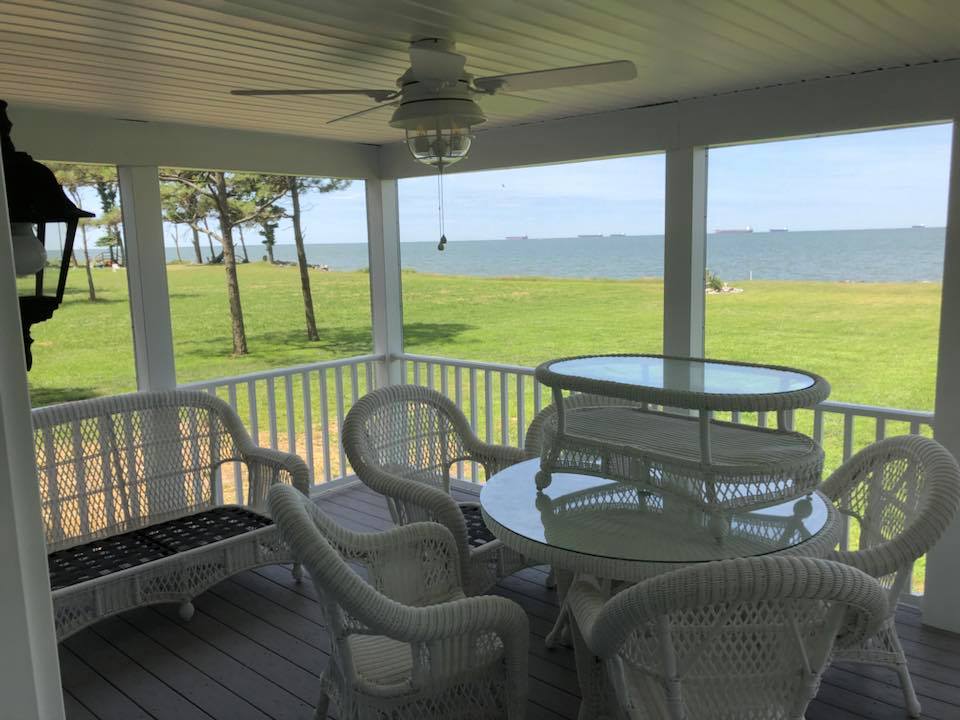 Covered porch with wicker furniture, including chairs and a glass-topped table, overlooking a green lawn and calm sea under a sunny sky. Relaxing vibe.