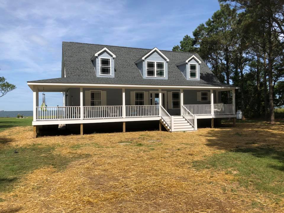 A large, two-story house with a gray roof, three dormer windows, and a wraparound porch. It's set in a grassy yard with tall trees, under a clear blue sky.