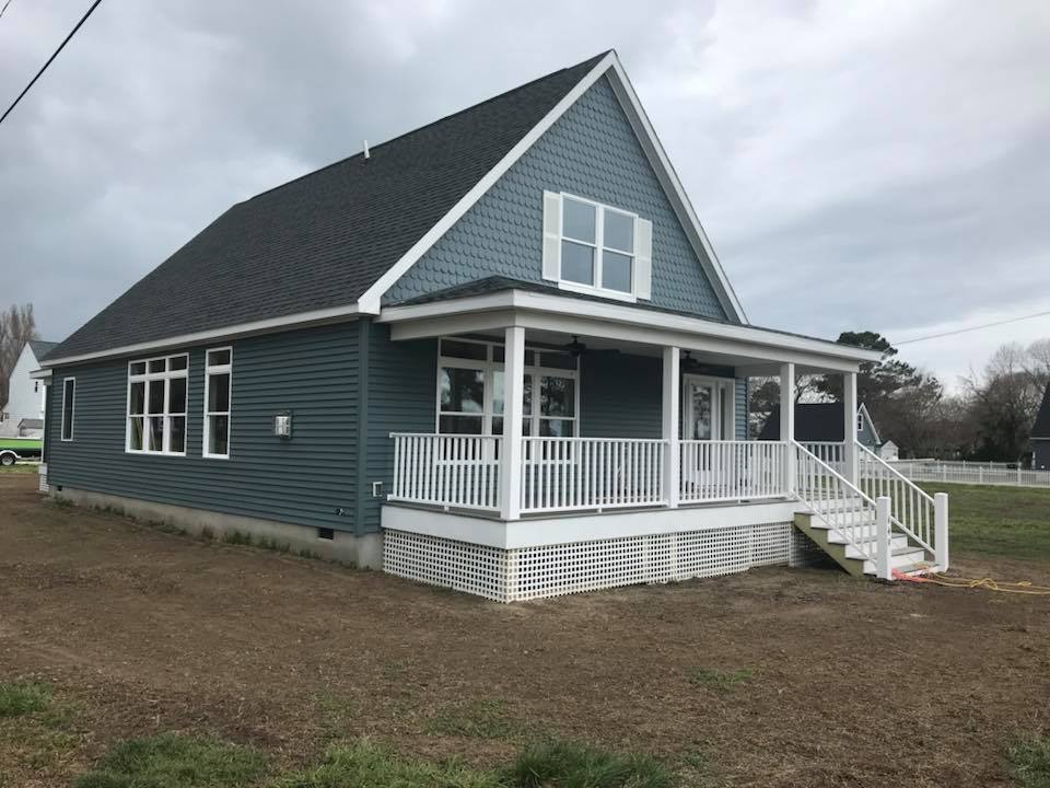A blue cottage-style house with a white front porch and A-frame roof sits against a cloudy sky. The house has multiple windows and a small lawn.